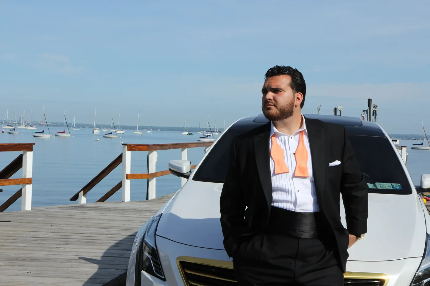 A singer in a tuxedo standing on a dock beside a car